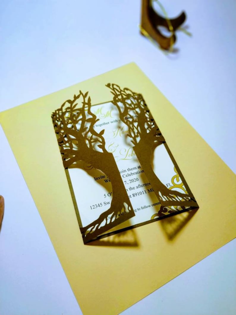 May include: A gold-colored wedding invitation with a laser-cut tree design. The invitation includes the text "Invite We" and event details. The invitation is on a gold background.