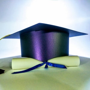 Puede incluir: Un gorro de graduaci&oacute;n negro con borla dorada sobre un diploma blanco enrollado atado con una cinta azul.