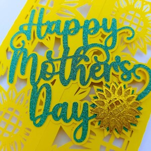 Op de afbeelding: Een gele kaart met een groene glitter-uitsparing van de woorden "Happy Mother's Day". De kaart is versierd met gele glitter-uitsparingen van zonnebloemen.