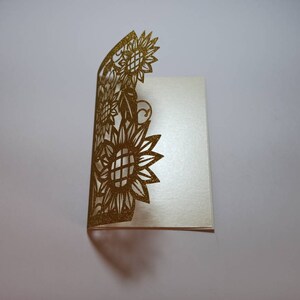 May include: A gold glitter card with a cut-out design of sunflowers. The card is folded in half and the design is on the front.
