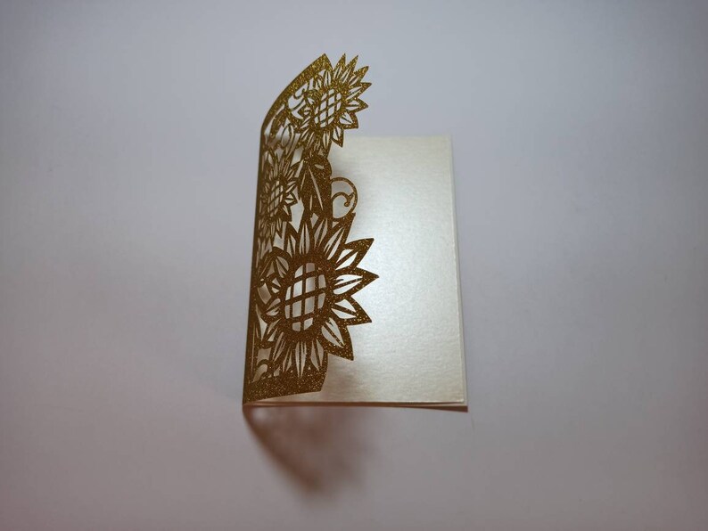 May include: A gold glitter card with a cut-out design of sunflowers. The card is folded in half and the design is on the front.