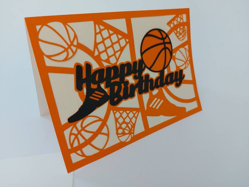 May include: Orange card with a white background and a black cut-out design of basketballs, a basketball hoop, and a shoe. The text "Happy Birthday" is cut out in black.