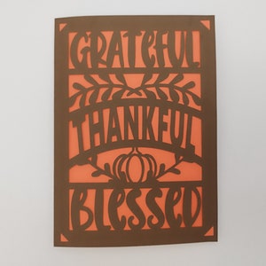 May include: A brown card with a cut-out design featuring the words "grateful", "thankful", and "blessed" in a stylized font. The background is a bright orange color.