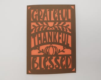 Könnte beinhalten: Eine braune Karte mit einem ausgeschnittenen Design, das die Wörter "grateful", "thankful" und "blessed" in einer stilisierten Schriftart zeigt. Der Hintergrund ist in leuchtendem Orange.