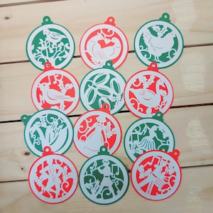 May include: A collection of Christmas ornaments with intricate cut-out designs. Each ornament features a red or green border with white interior images of birds, flowers, and figures. The ornaments are arranged on a light brown wooden surface.