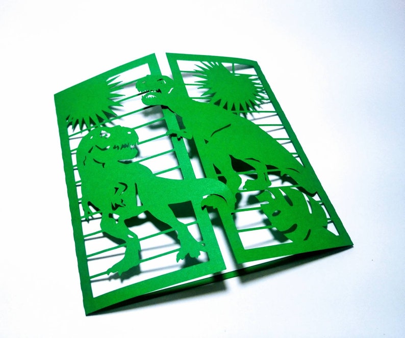 May include: Green paper card with a cut-out design of two dinosaurs in a jungle setting. The card is folded in half and the dinosaurs are visible on both sides.