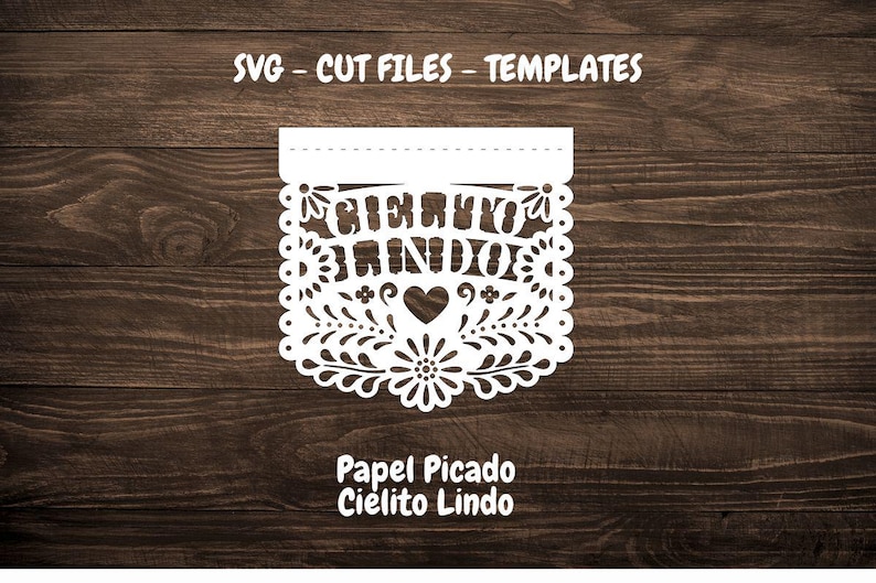 May include: White paper cut-out banner with a floral design and the text "Cielito Lindo" in Spanish. The banner is on a brown wooden background. The text "Papel Picado Cielito Lindo" is below the banner.