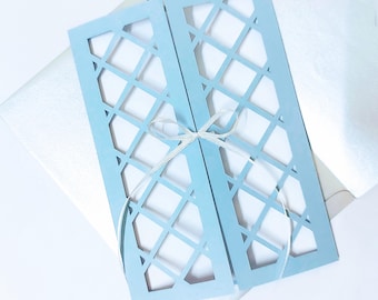 May include: A light blue invitation card with a lattice design, tied with a white ribbon. The card is open, revealing a white interior. It rests on a white envelope, suggesting a wedding or special event theme.