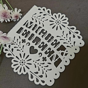 May include: White paper cut-out with the words "Mi Querido Amor" in a decorative font, surrounded by floral and heart designs. The edges are scalloped. A few white and purple flowers are in the upper left corner.