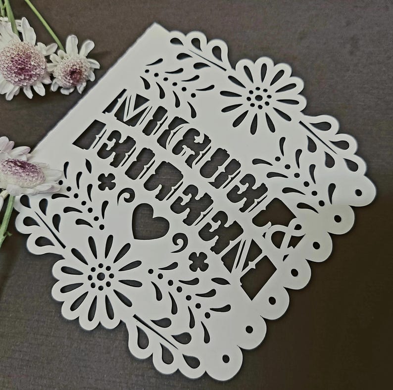 May include: White paper cut-out with the words "Mi Querido Amor" in a decorative font, surrounded by floral and heart designs. The edges are scalloped. A few white and purple flowers are in the upper left corner.