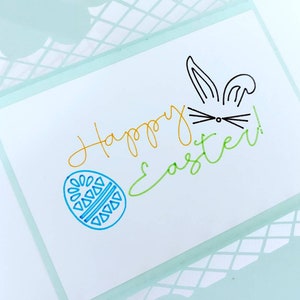 May include: A white card with the words "Happy Easter!" written in green, yellow, and blue. A blue egg with a geometric pattern is drawn on the left side of the card. A black bunny with ears and whiskers is drawn on the right side of the card.
