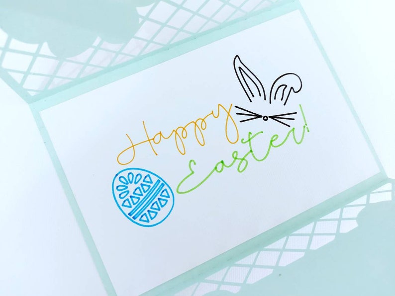 May include: A white card with the words "Happy Easter!" written in green, yellow, and blue. A blue egg with a geometric pattern is drawn on the left side of the card. A black bunny with ears and whiskers is drawn on the right side of the card.