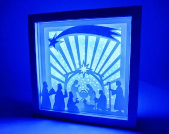 May include: A white wooden frame with a blue light shining through a paper cut-out of the nativity scene. The scene includes a star, a manger, and figures of Mary, Joseph, and the baby Jesus.
