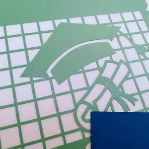 Op de afbeelding: Een groene papieren sjabloon met een ontwerp van een graduatiecap en een diploma. De sjabloon bevindt zich op een witte rasterachtergrond.