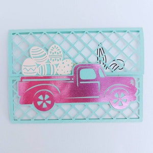 May include: A pink pickup truck with a white bed filled with colourful Easter eggs. The truck is cut out of paper and is sitting on a light blue background.
