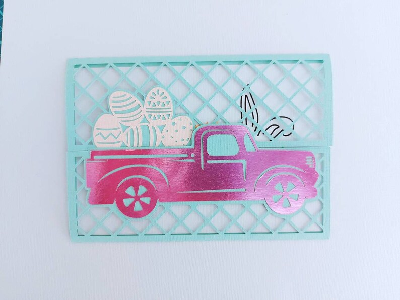 May include: A pink pickup truck with a white bed filled with colourful Easter eggs. The truck is cut out of paper and is sitting on a light blue background.