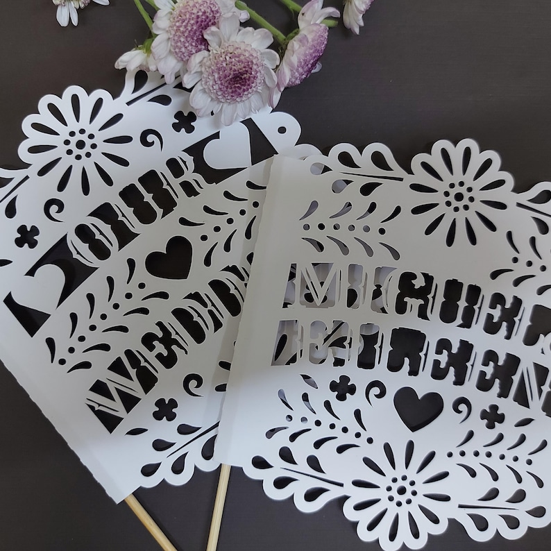 May include: White paper flags with intricate cut-out designs, featuring floral and heart motifs, and the words "OUR WEDDING" and "MICHELLE & BRENDAN". The flags are on wooden sticks, with purple flowers in the background.
