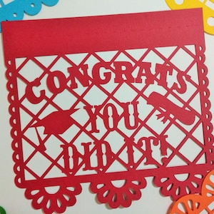 Op de afbeelding: Rode papieren banner met een wit uitgesneden ontwerp met de tekst "Congrats You Did It!" De banner heeft een gegolfde rand en een decoratief patroon.