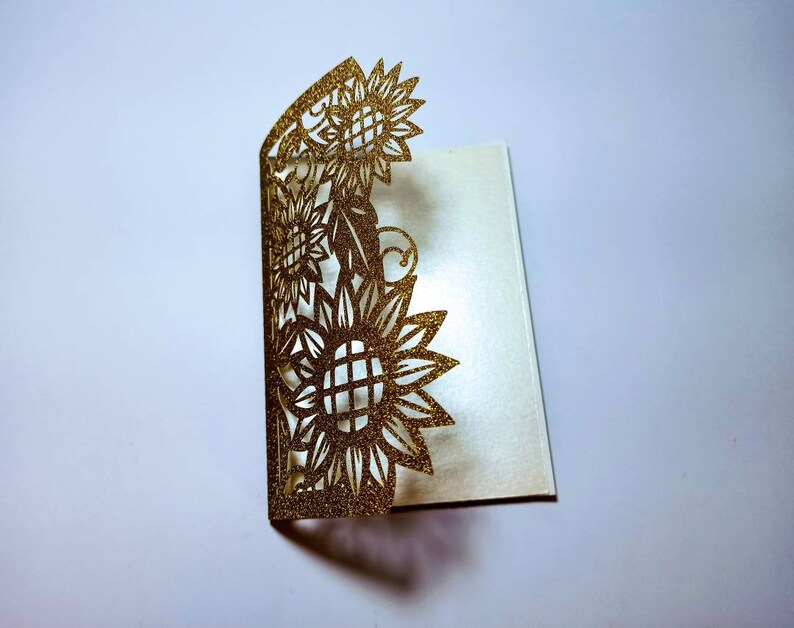 May include: Gold glitter laser cut card with a sunflower design. The card is folded in half and the design is on the front.