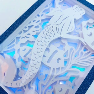 May include: A white paper cutout of a mermaid with a fish and seaweed in a blue frame. The mermaid is in a sitting position with her tail curled around her.