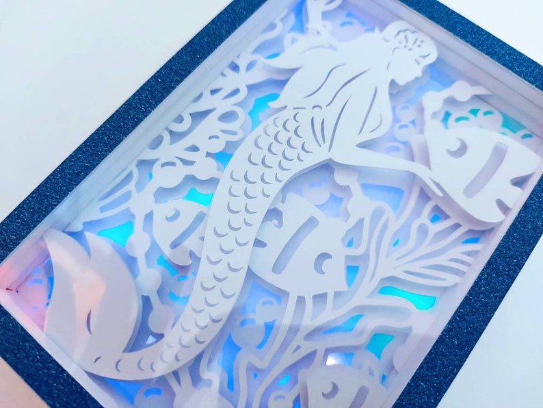 May include: A white paper cutout of a mermaid with a fish and seaweed in a blue frame. The mermaid is in a sitting position with her tail curled around her.