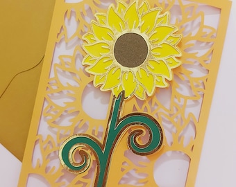 Può includere: Un biglietto di carta giallo e verde con un design ritagliato a forma di girasole. Il girasole ha un centro marrone e petali gialli. Il biglietto è piegato a metà e ha una busta dorata.