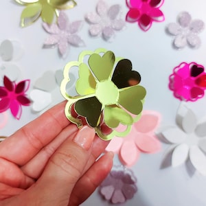 May include: A gold-coloured, reflective, heart-shaped flower cutout held in the foreground, surrounded by various scattered paper flower shapes in shades of pink, white, light purple, and magenta with glittery and metallic finishes.