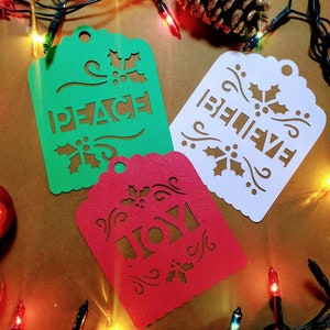 May include: Three paper Christmas ornaments with cut-out designs. The ornaments are green, white, and red. The green ornament says "PEACE", the white ornament says "BELIEVE", and the red ornament says "JOY".