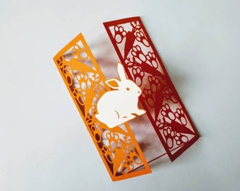 May include: A white paper cut-out of a rabbit is layered over a red and orange paper card with a cut-out design of carrots and paw prints.