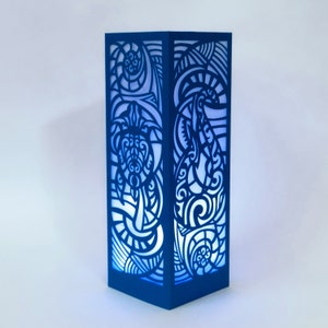 Puede incluir: Una lámpara azul rectangular con intrincados diseños recortados. Los lados de la lámpara presentan un patrón repetido de líneas y formas arremolinadas, con un diseño de tortuga. La lámpara emite un suave brillo azul, creando un efecto de iluminación decorativo.