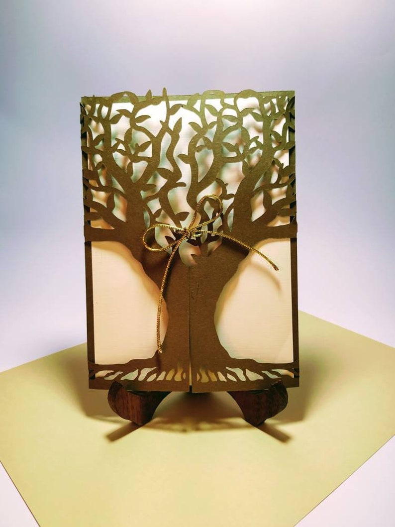 May include: A laser-cut olive-green card with a tree design. The card is held open by a small wooden stand, revealing a cream-colored interior. A gold-colored string is tied around the card. The card is on a light yellow surface.