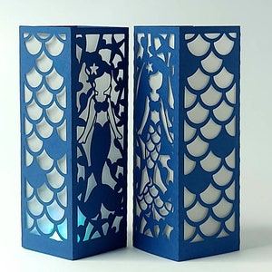 Puede incluir: Dos faroles de papel azul con un dise&ntilde;o de silueta de sirena. Los faroles tienen un fondo blanco y est&aacute;n decorados con un patr&oacute;n de escamas.