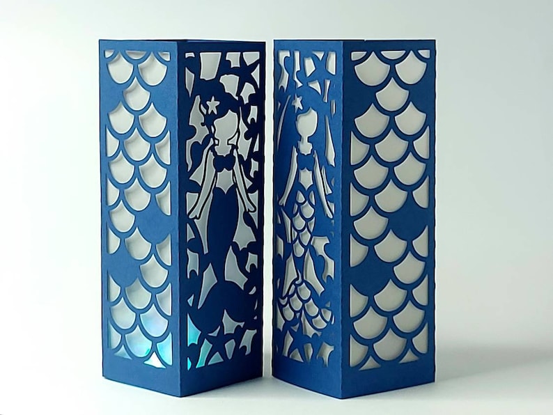 Puede incluir: Dos faroles de papel azul con un dise&ntilde;o de silueta de sirena. Los faroles tienen un fondo blanco y est&aacute;n decorados con un patr&oacute;n de escamas.
