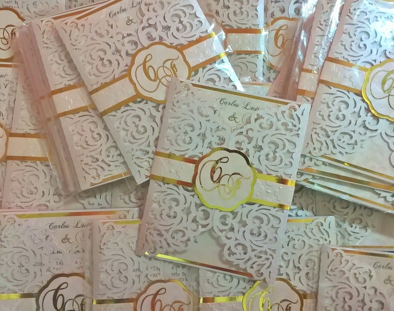 May include: A stack of white and gold wedding invitations with a delicate lace design. The invitations feature the names "Carlos Luis" and a gold monogram.