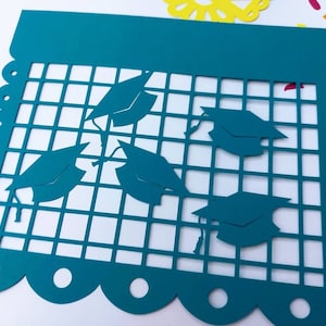 Op de afbeelding: Turquoise blauwe papieren banner met een gegolfde rand en een rasterpatroon. De banner heeft vijf uitgesneden afstudeerhoeden.