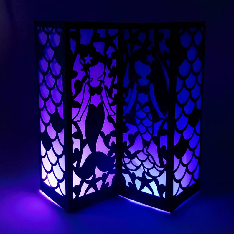 Puede incluir: Dos linternas de papel negras con un dise&ntilde;o de silueta de sirena. Las linternas est&aacute;n iluminadas desde el interior, proyectando un brillo morado y azul.