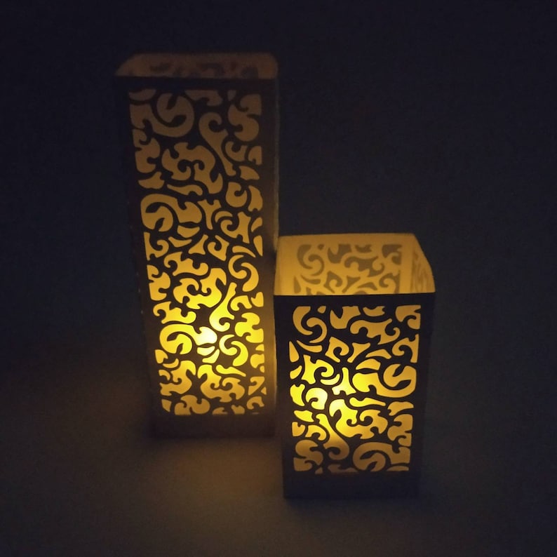May include: Two white paper lanterns with intricate cut-out designs, illuminated from within by a warm yellow light.