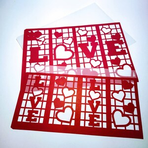 May include: Red paper cutout of the word "LOVE" with a grid pattern and small hearts. The cutout is layered on top of a white sheet of paper.