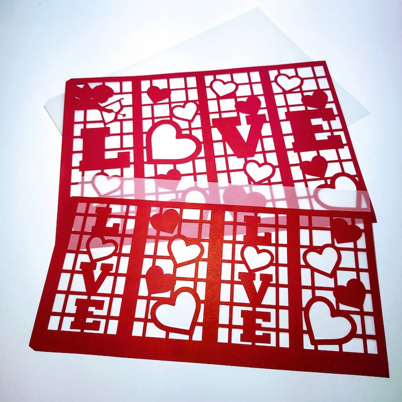 May include: Red paper cutout of the word "LOVE" with a grid pattern and small hearts. The cutout is layered on top of a white sheet of paper.