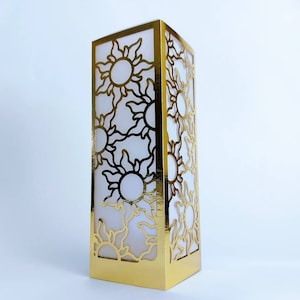 May include: Gold-colored decorative lantern with a cutout design of multiple suns. The lantern is made of paper and has a white interior.