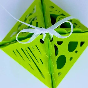 May include: A lime green paper gift box with a white ribbon. The box has a cut-out design with geometric shapes.