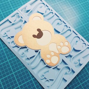 May include: A light blue card with a cutout design of hearts and swirls. A gold teddy bear with brown eyes and white paws is layered on top of the card.