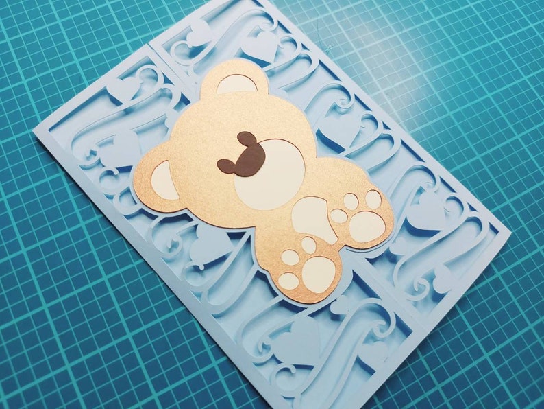 May include: A light blue card with a cutout design of hearts and swirls. A gold teddy bear with brown eyes and white paws is layered on top of the card.