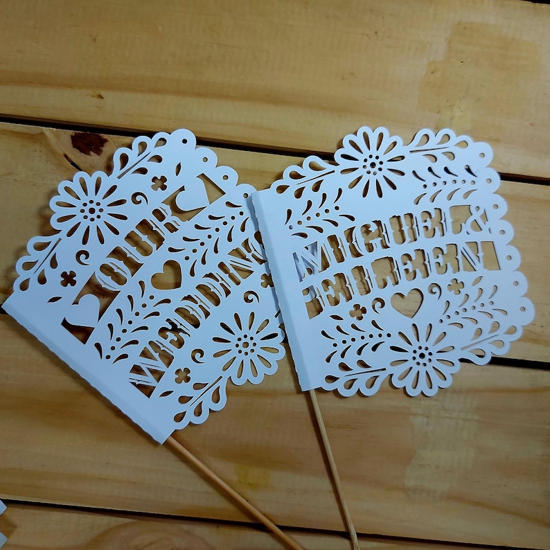 May include: Two white, laser-cut wedding cake toppers. One reads "OUR WEDDING" and the other reads "MICHELLE & BENJAMIN". Both have floral and heart cutouts and are mounted on wooden sticks, perfect for a wedding cake.