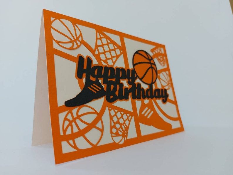 May include: Orange card with a white interior and a black cut-out design of basketballs, sneakers, and a basketball hoop. The text "Happy Birthday" is cut out in black.