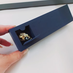 May include: A small, gold-colored, decorative object with a black and white design is inside a dark blue box with a cutout.