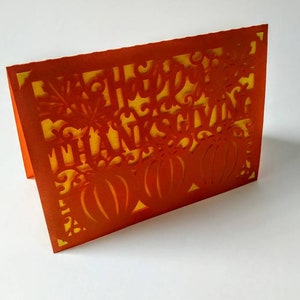 Puede incluir: Tarjeta naranja con fondo amarillo y un dise&ntilde;o recortado de calabazas y las palabras "Feliz D&iacute;a de Acci&oacute;n de Gracias".