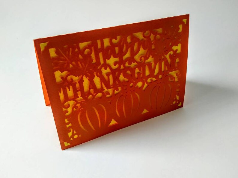 Puede incluir: Tarjeta naranja con fondo amarillo y un dise&ntilde;o recortado de calabazas y las palabras "Feliz D&iacute;a de Acci&oacute;n de Gracias".