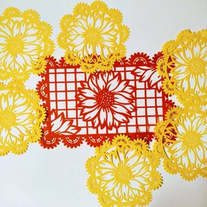 Puede incluir: Un conjunto de seis recortes de papel amarillo de girasoles con dise&ntilde;os intrincados. Los recortes est&aacute;n dispuestos en un c&iacute;rculo alrededor de un recorte de papel rojo de un patr&oacute;n de cuadr&iacute;cula con un dise&ntilde;o de girasol en el centro.