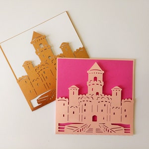 May include: Two paper cards with a castle design cut out. The top card is white with a gold castle design. The bottom card is pink with a pink castle design.